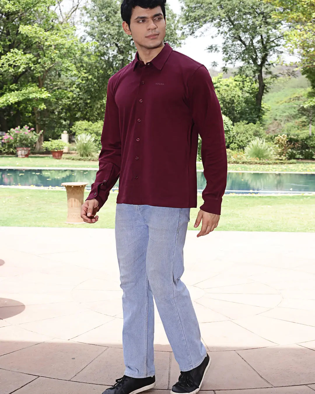 Man wearing a maroon shirt and light blue jeans standing outdoors with greenery in the background