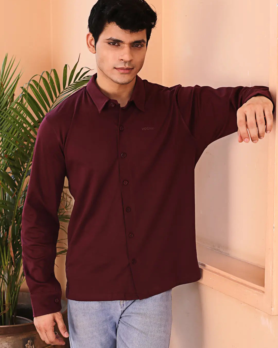 Man wearing a maroon shirt and light blue jeans standing indoors with a plant in the background