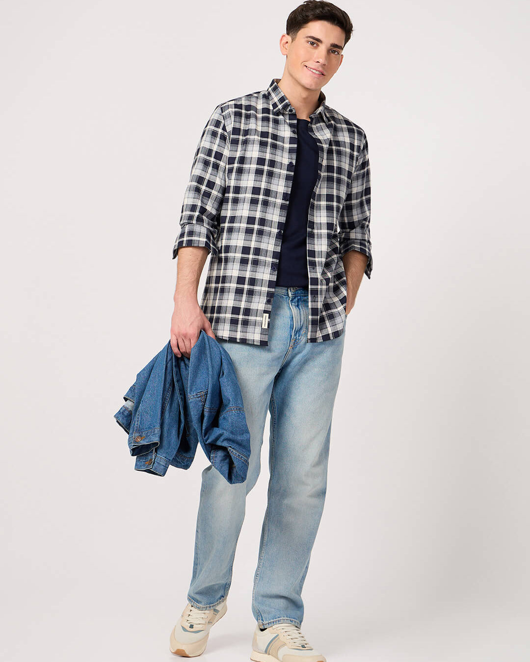 Man wearing a plaid shirt, blue jeans, and holding a denim jacket on a white background