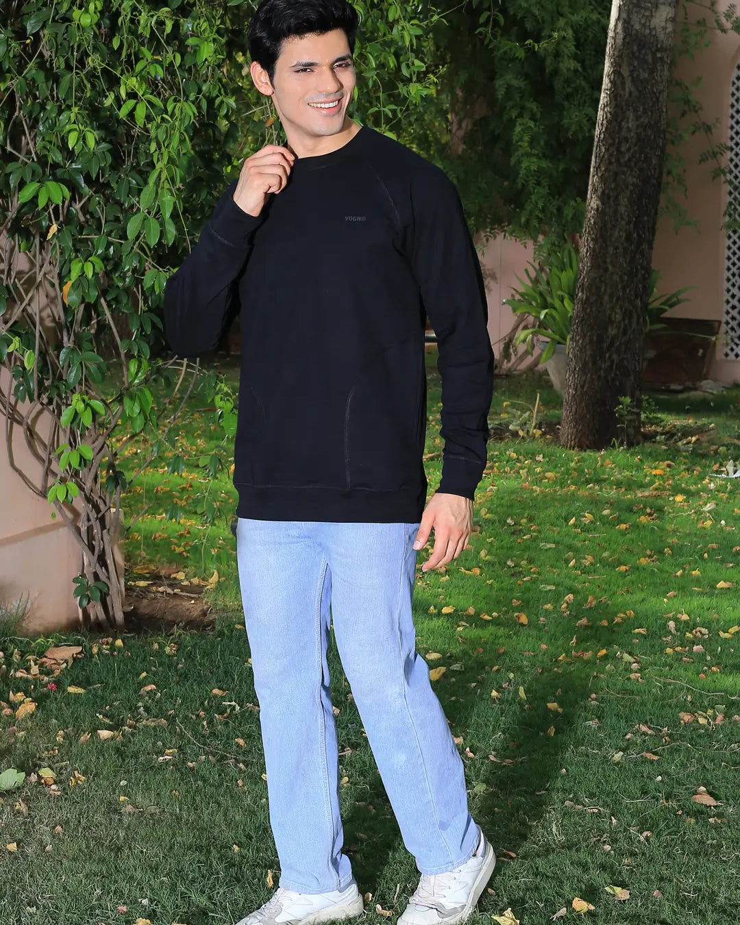 Man wearing a black sweatshirt and light blue jeans standing outdoors with greenery in the background