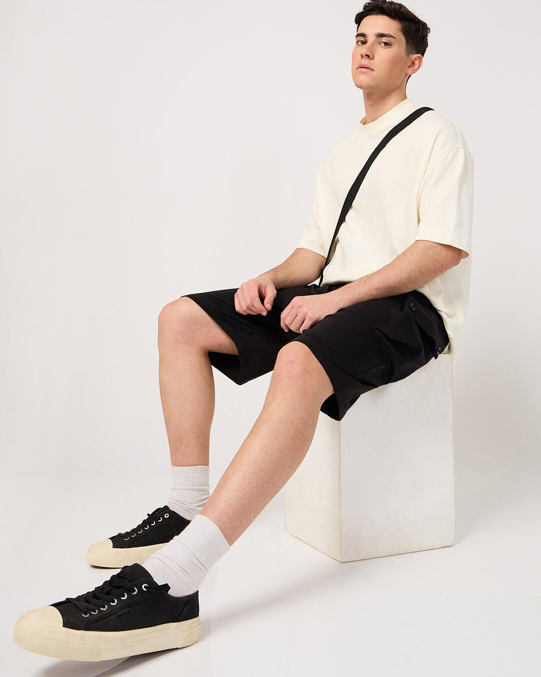 Person sitting on a white chair wearing a white shirt, black cargo shorts, and black sneakers with white socks.