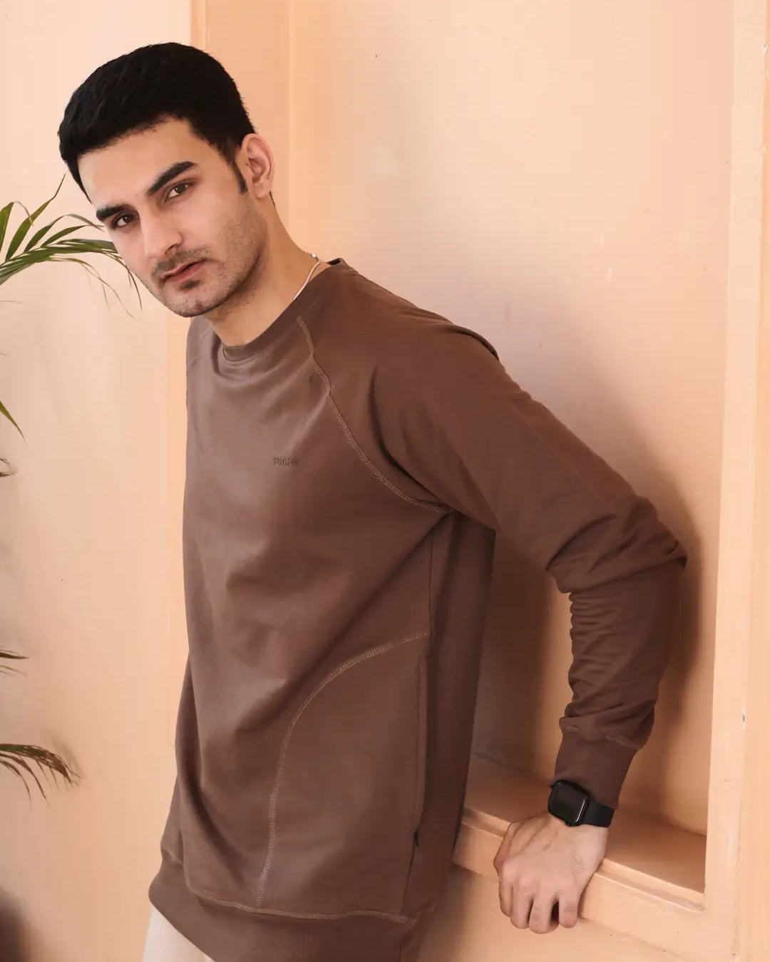 Man wearing a brown sweatshirt standing against a beige wall