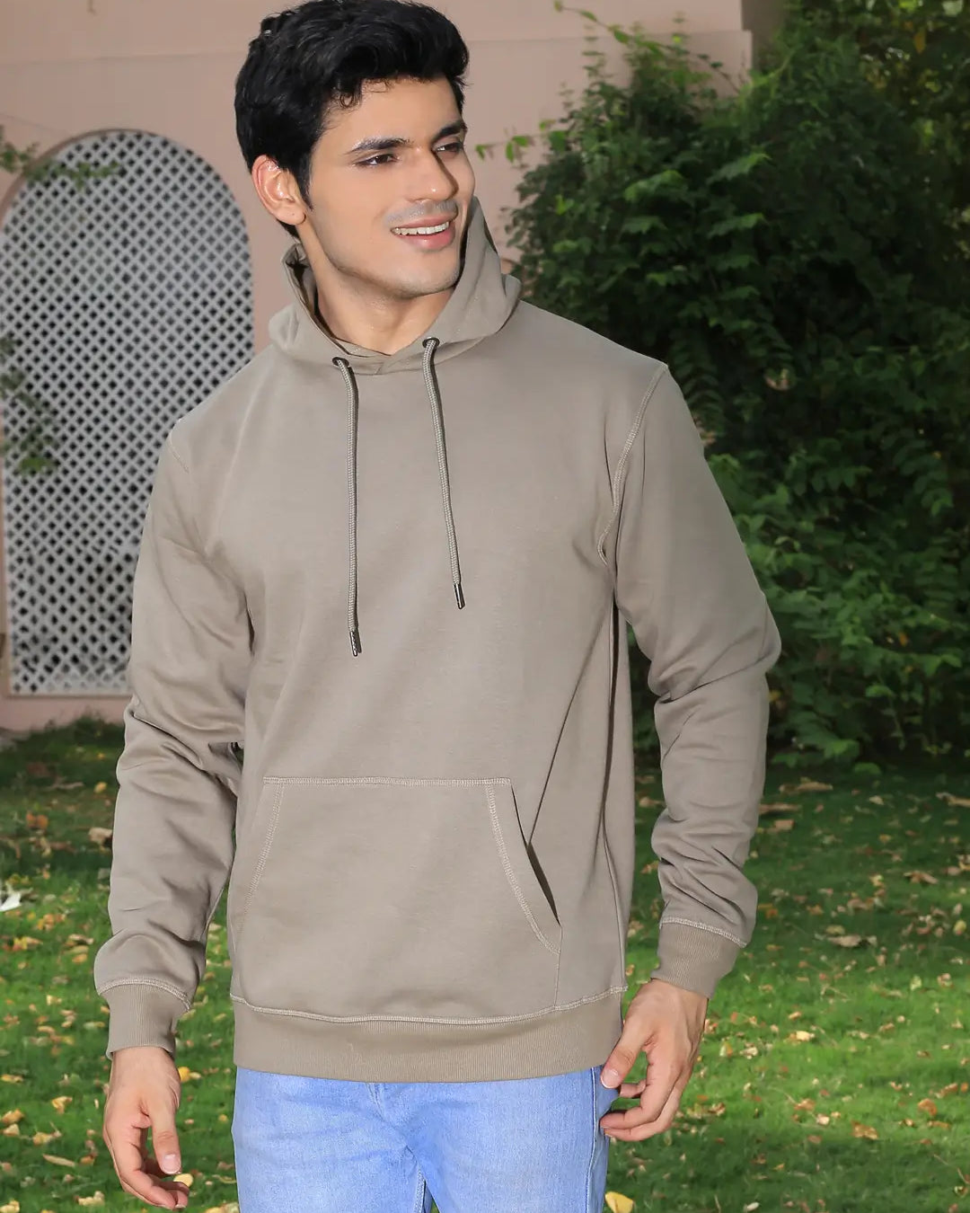 Man wearing a grey hoodie and blue jeans standing outdoors with greenery in the background