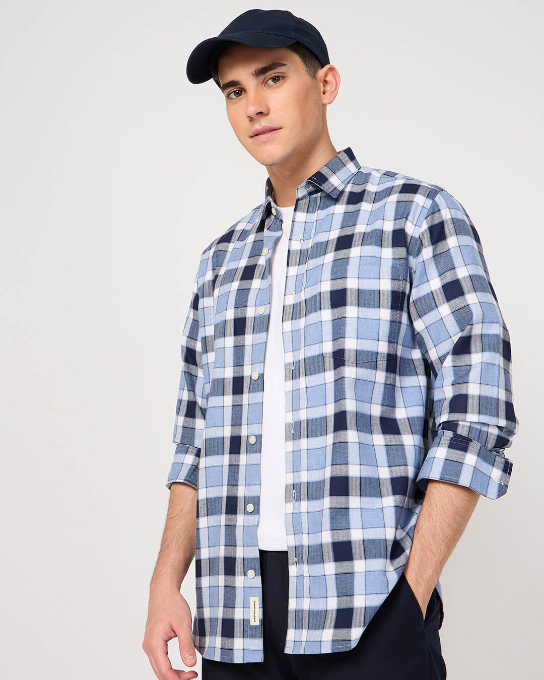Man wearing a blue and white checkered shirt with rolled-up sleeves and a navy cap on a light gray background