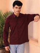 Man wearing a maroon shirt and light blue jeans standing indoors with a plant in the background